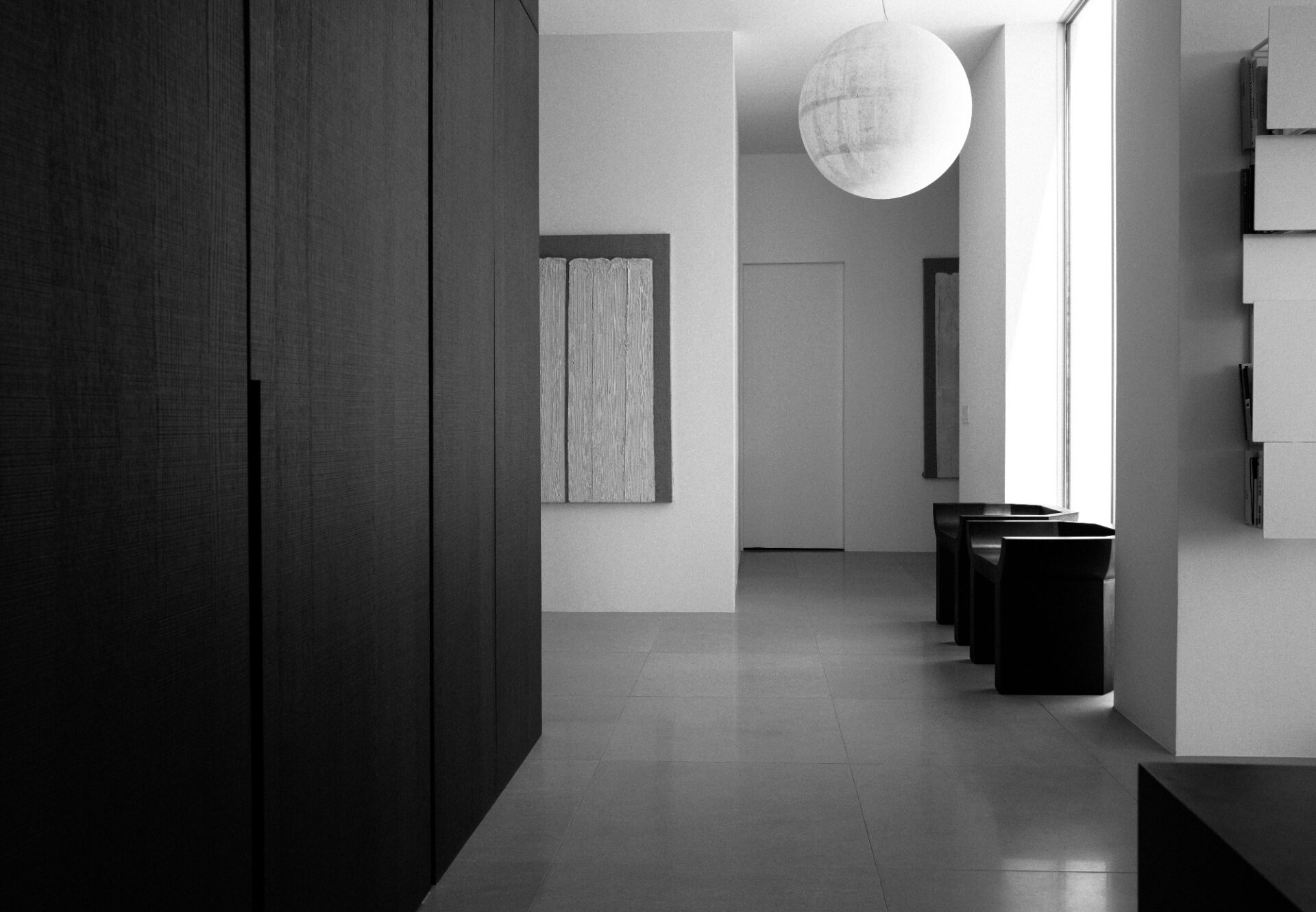 Minimalist hallway with dark wood cabinetry on left, large window on right, black stools, and modern pendant light.