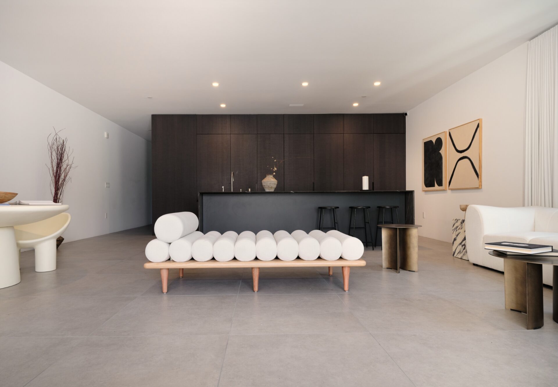Open-plan living space with sculptural daybed, dark kitchen cabinetry, and modern minimalist furnishings.