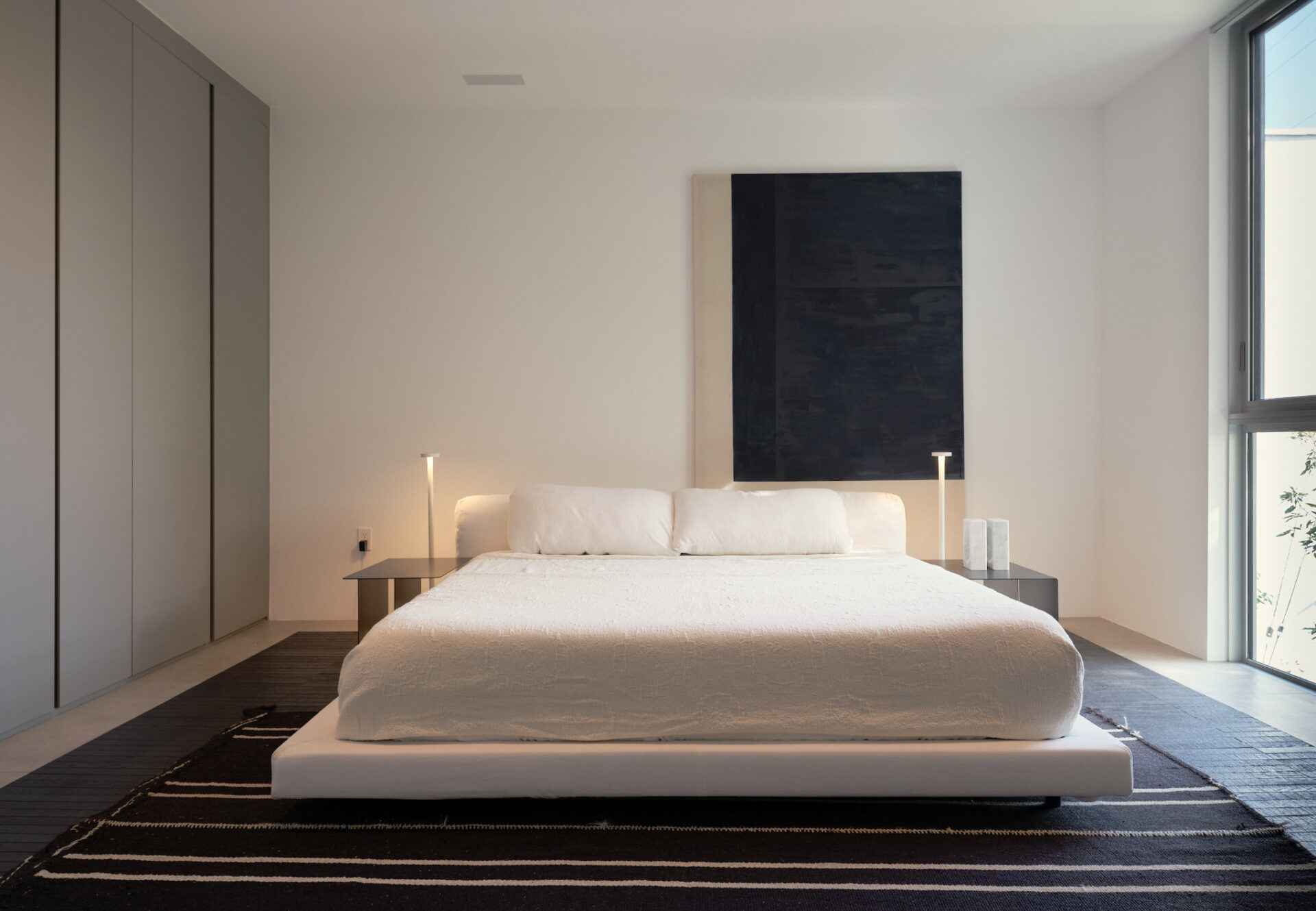 Minimalist bedroom with tall ceilings, built-in wardrobe, abstract artwork, and soft natural light from a large window.