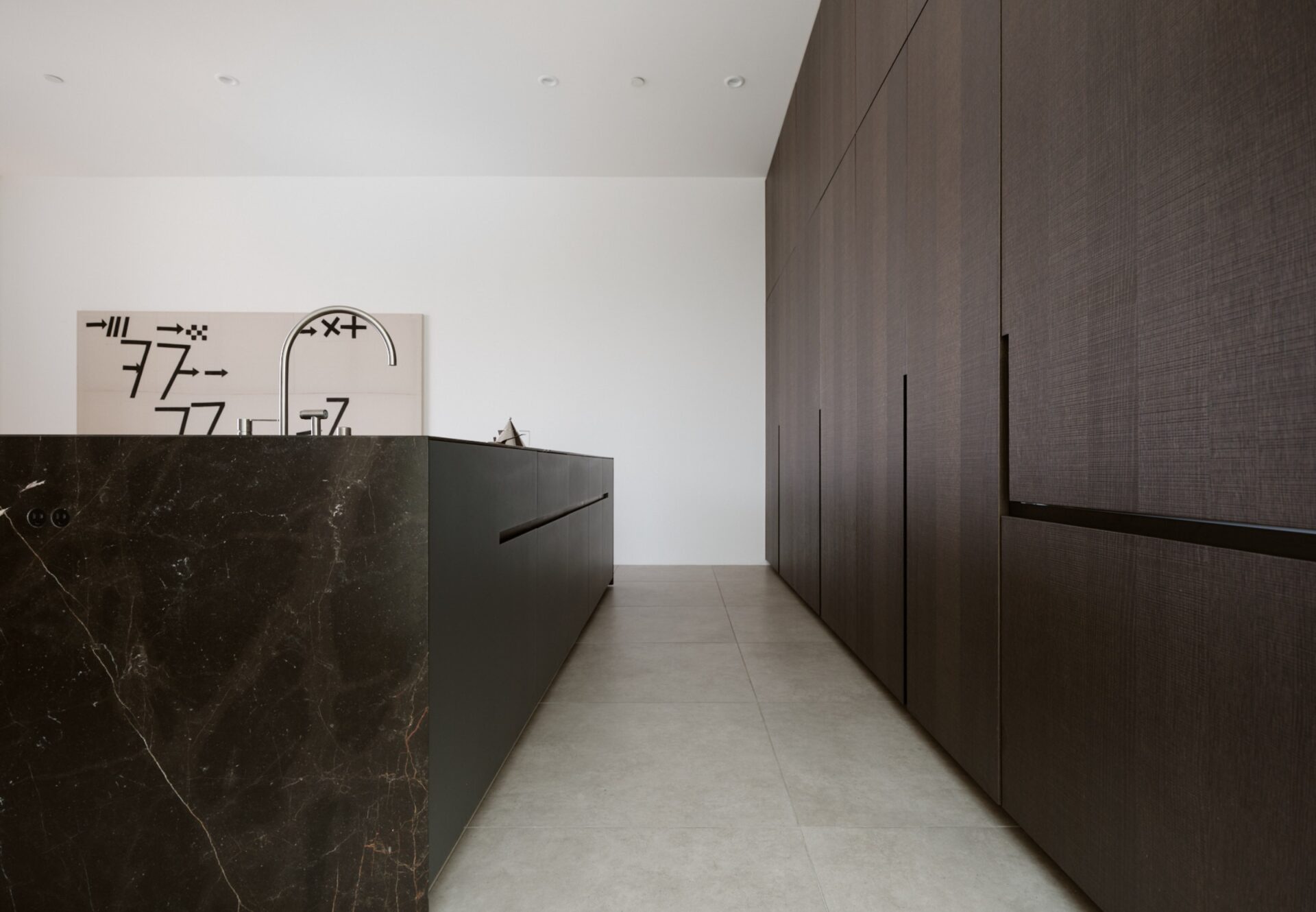 Profile view of a minimalist kitchen featuring floor-to-ceiling dark oak cabinetry, a central marble island, and high ceilings.