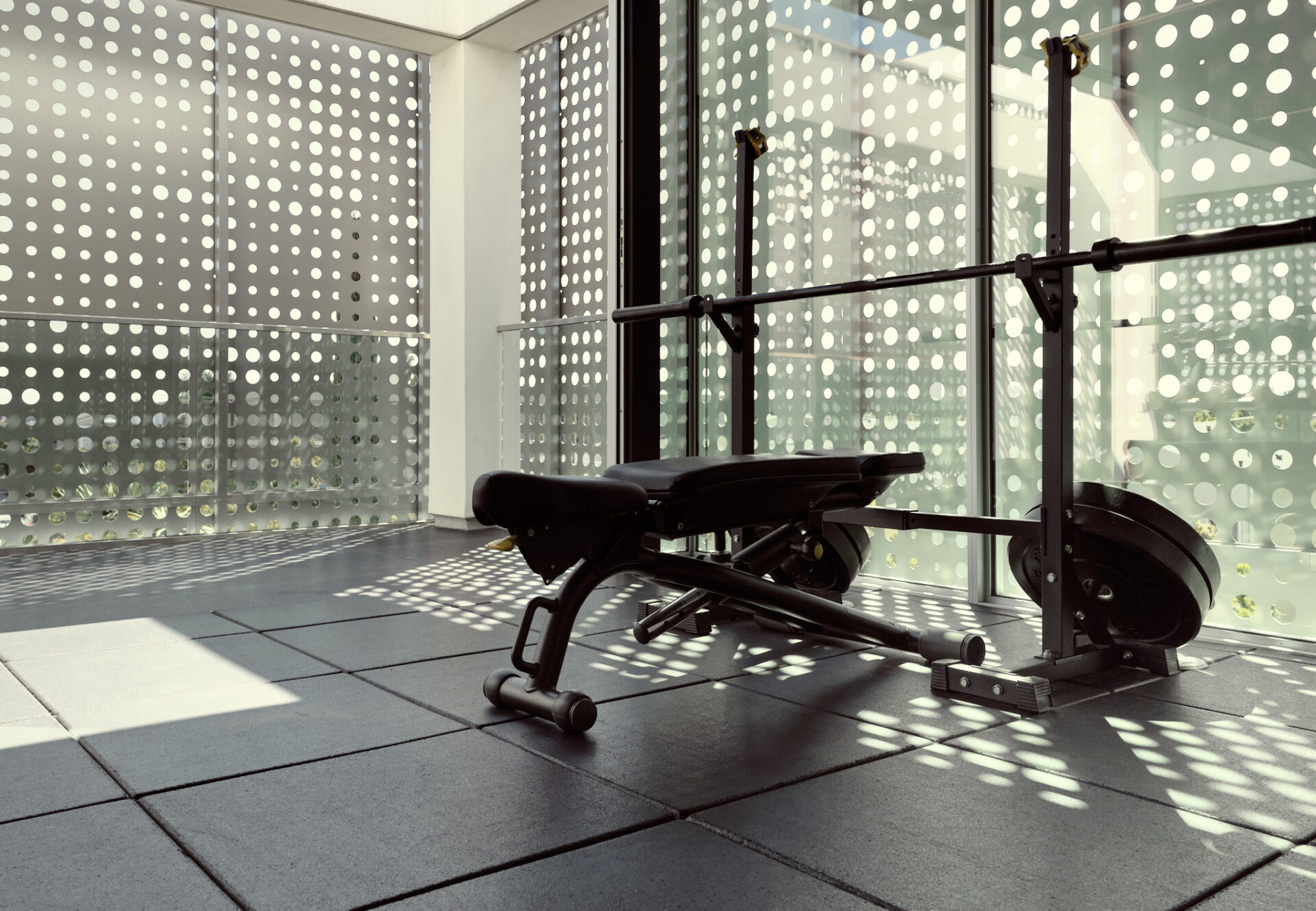 Penthouse fitness studio with weight bench and barbell beside perforated metal screens casting patterned light.