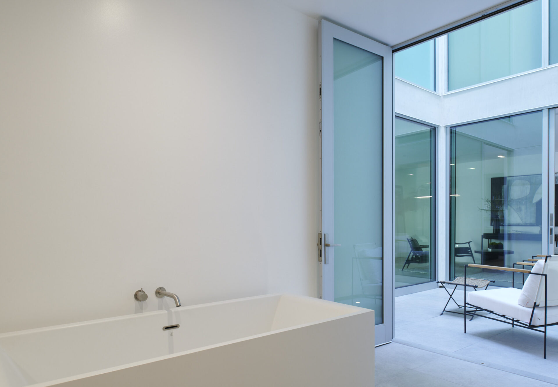 Freestanding bathtub beside glass door opening to a private furnished atrium in a bright, indoor-outdoor en suite bath.
