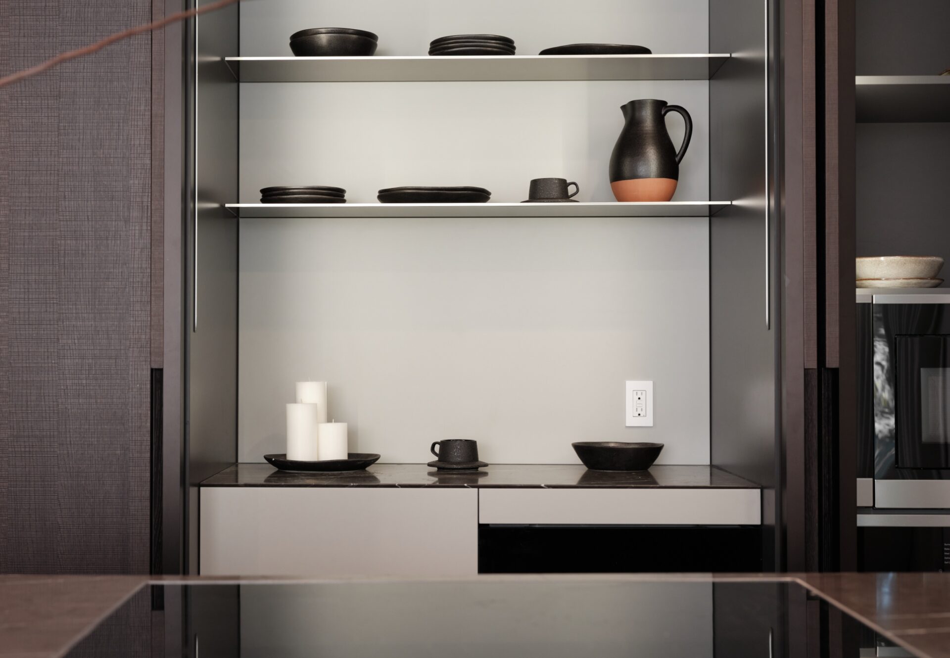Detail of minimalist kitchen shelving with ceramic dishware illuminated by recessed lighting, near electric stovetop and dark cabinetry.