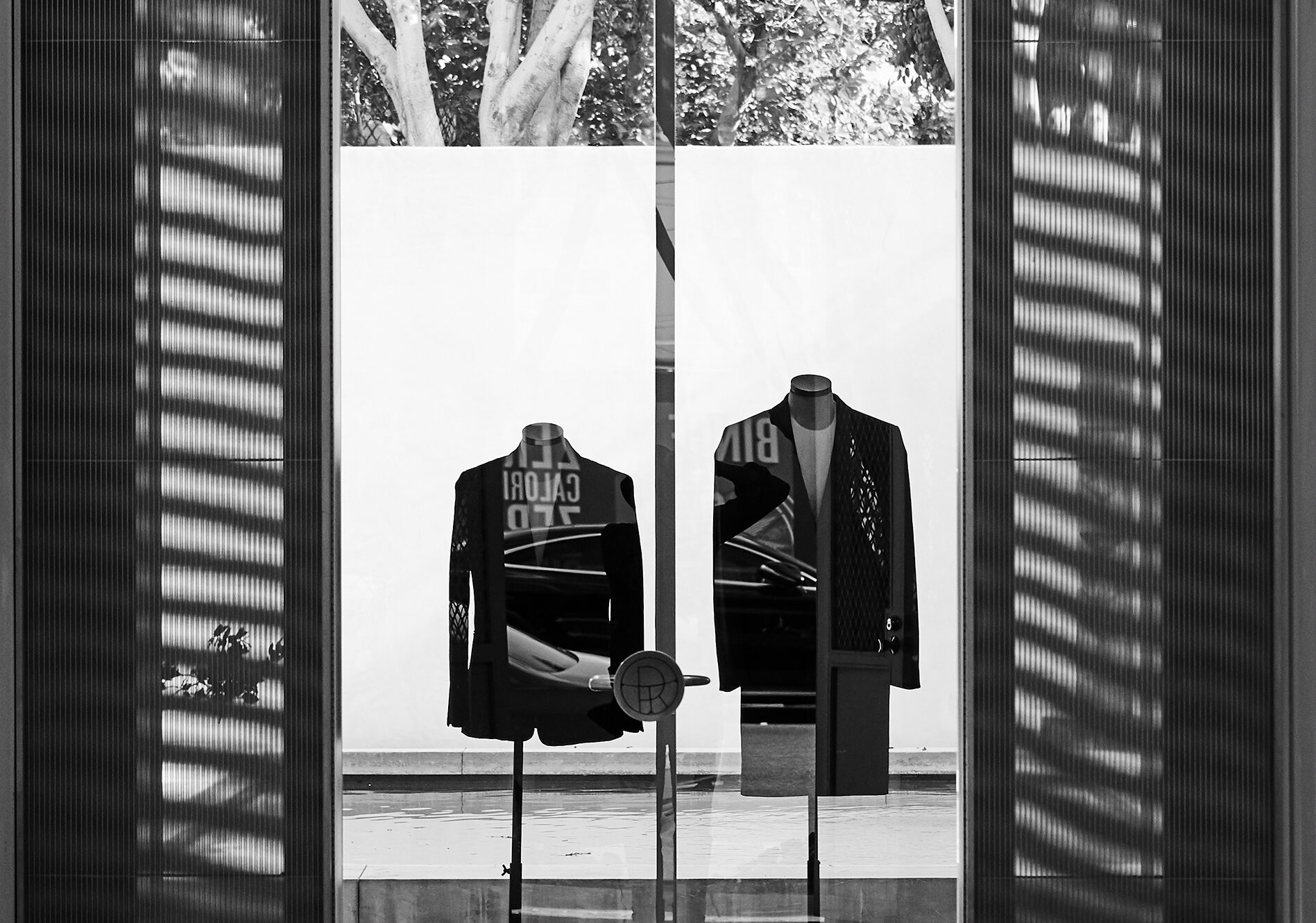 View through open doorway showing two mannequins dressed in dark jackets, with window shadows casting lines on walls.