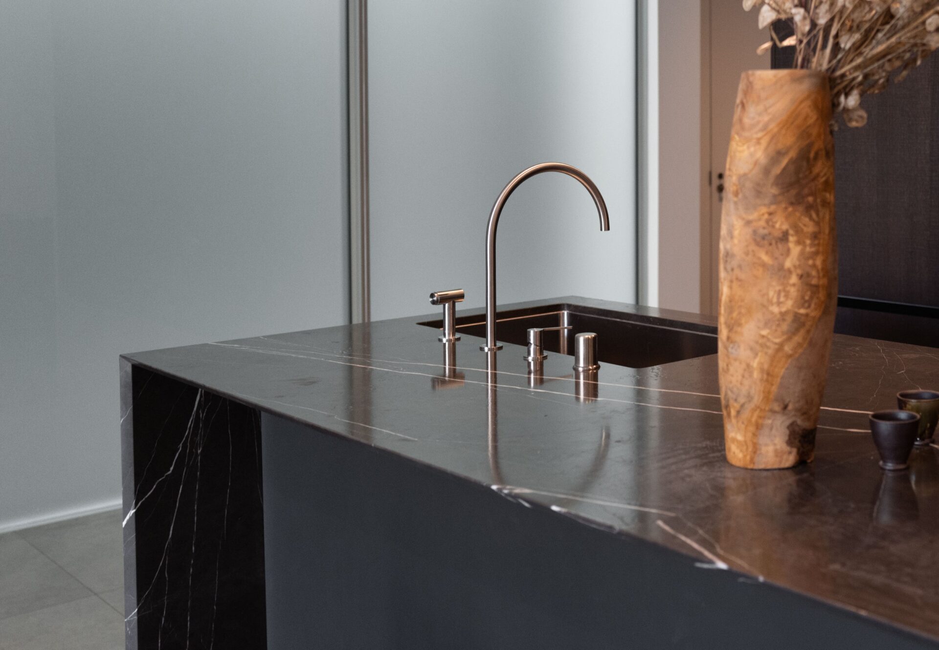 Close-up of a marble kitchen island featuring a waterfall countertop, modern faucet fixtures, and a tall stone vase accent.