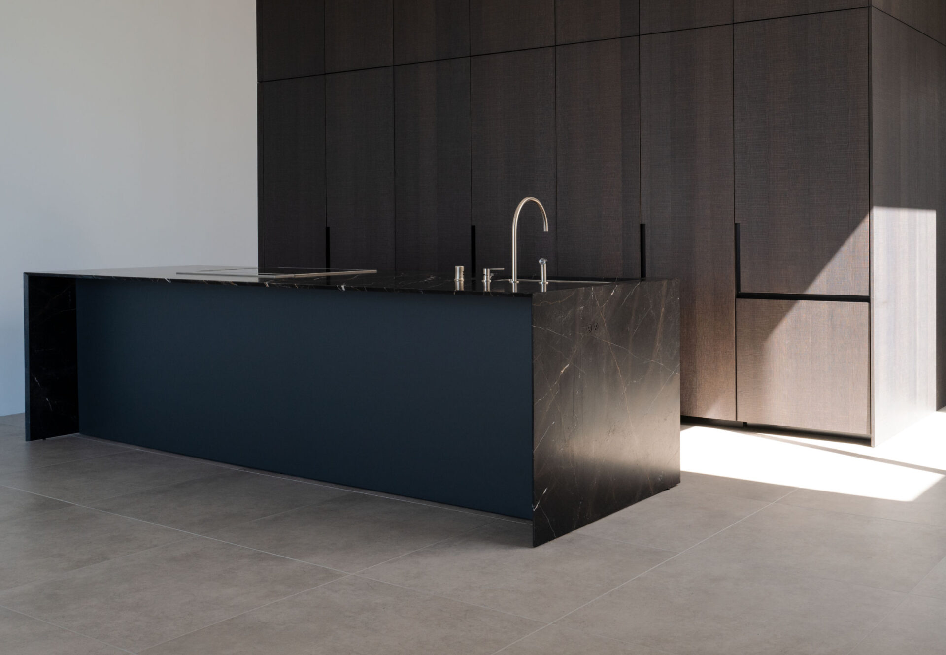 Minimalist kitchen with dark oak cabinetry, black marble island, and natural light casting geometric shadows on the floor.