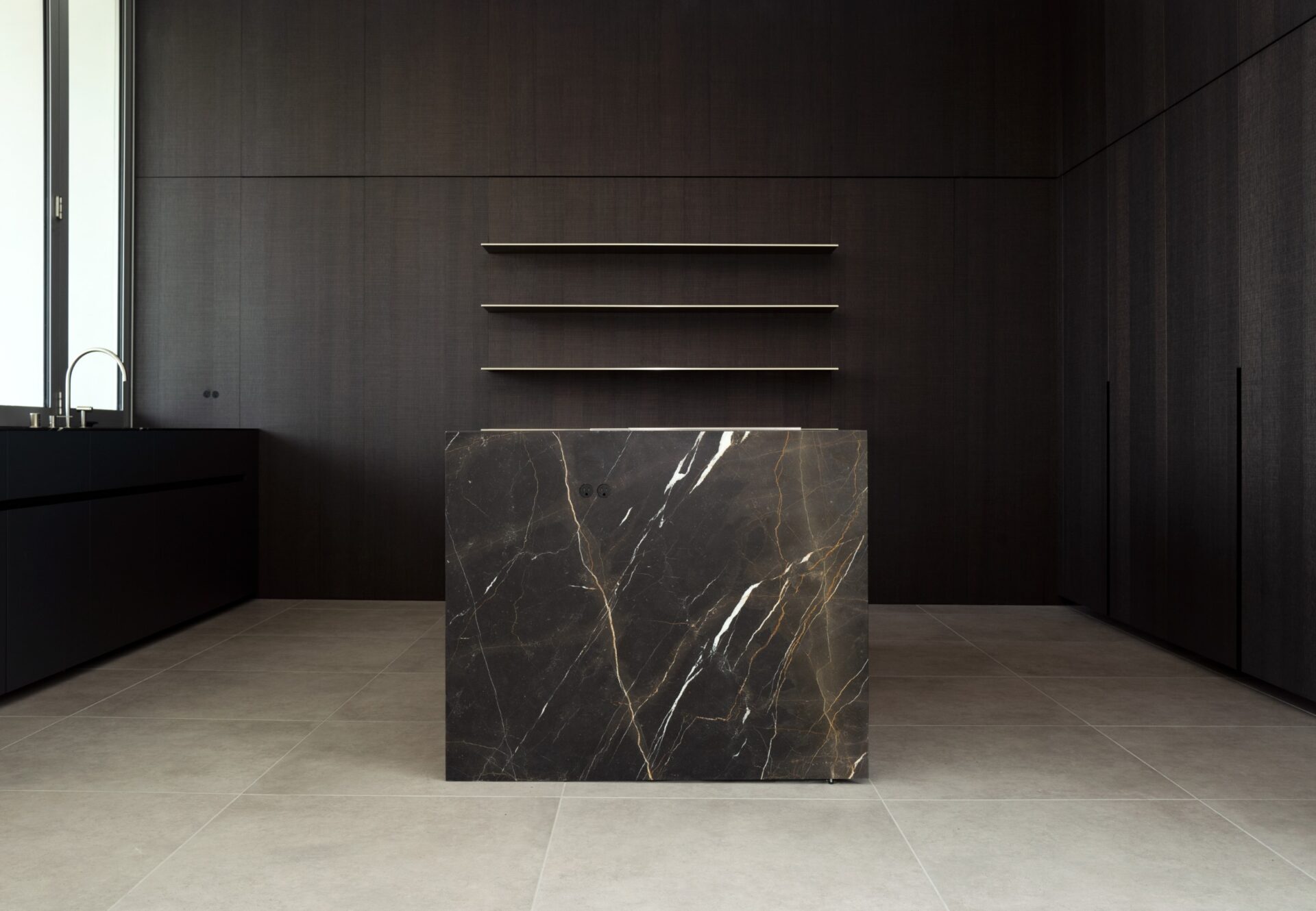 Minimalist kitchen with dark oak cabinetry, bold marble island, neutral stone flooring, and open shelving.