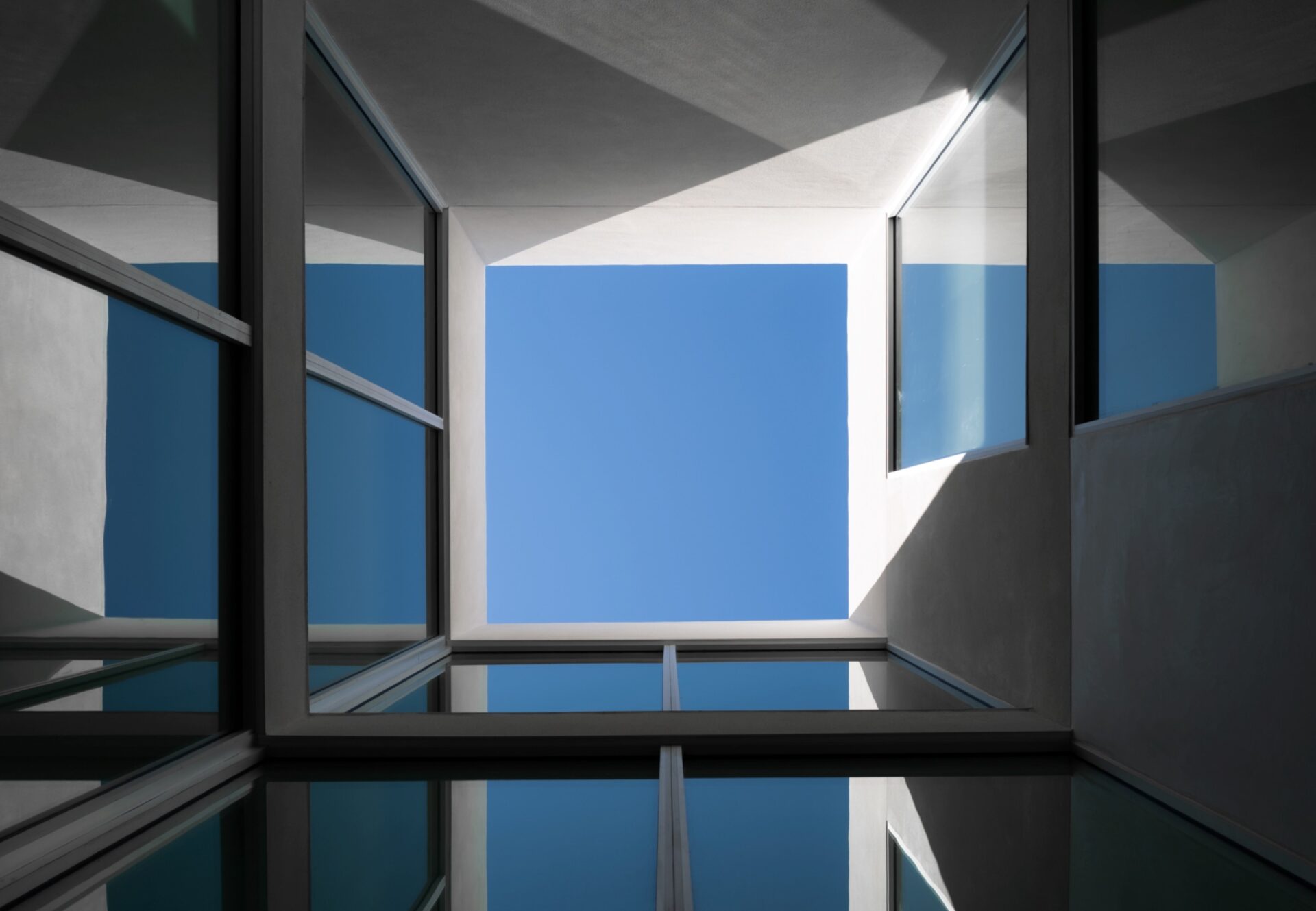 Upward view of a modern geometric atrium with glass walls reflecting a clear blue sky.