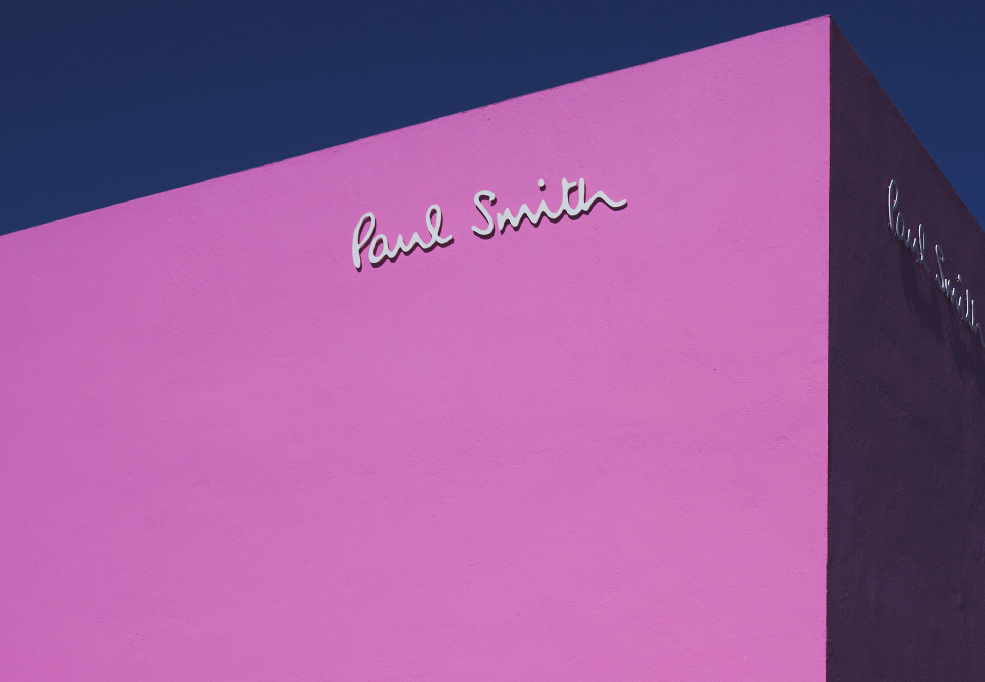Bright pink exterior wall of Paul Smith building with white signature logo against clear blue sky.
