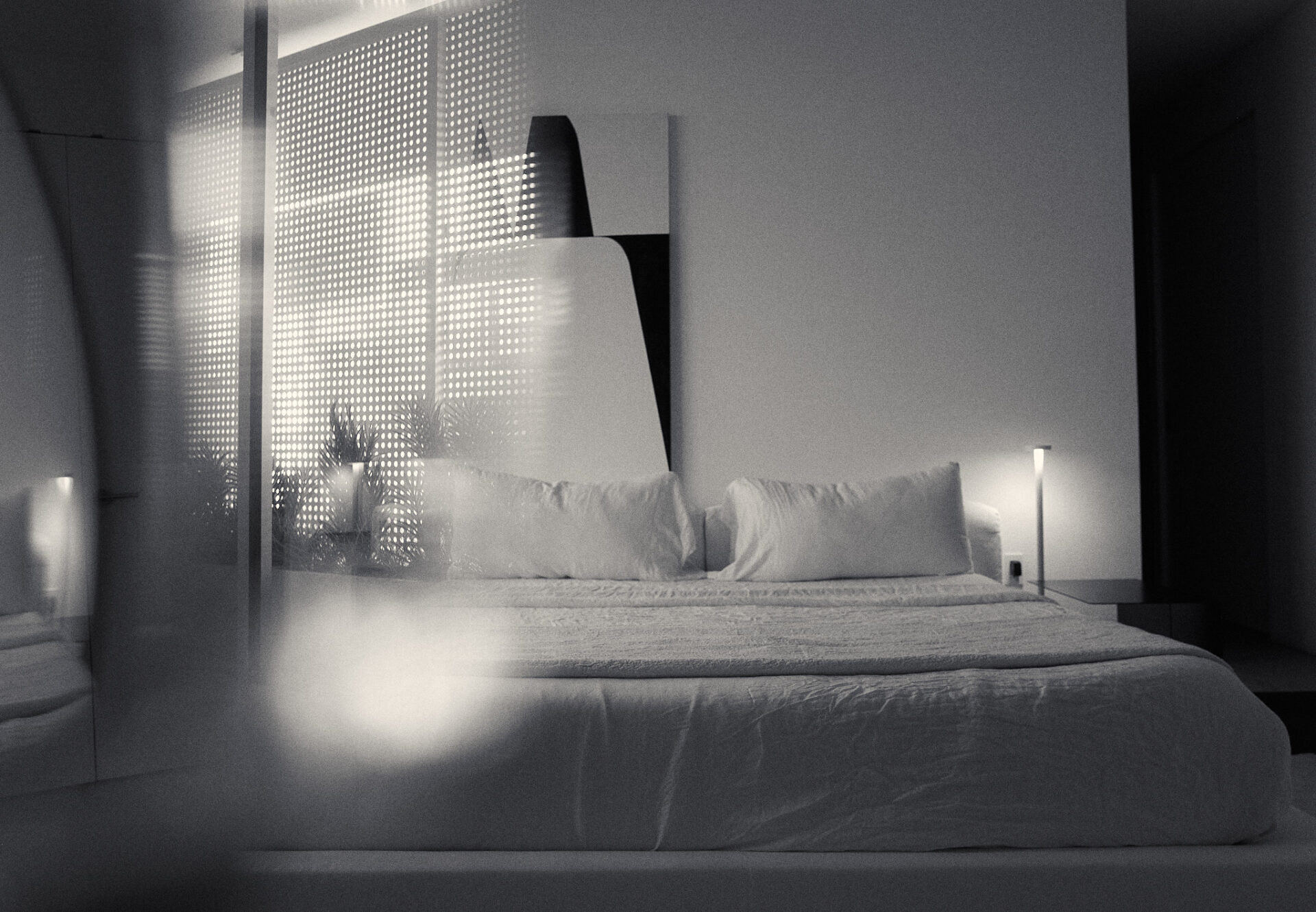 Primary bedroom with low-profile bed and abstract art, softly lit and partially obscured by perforated veil privacy screen.