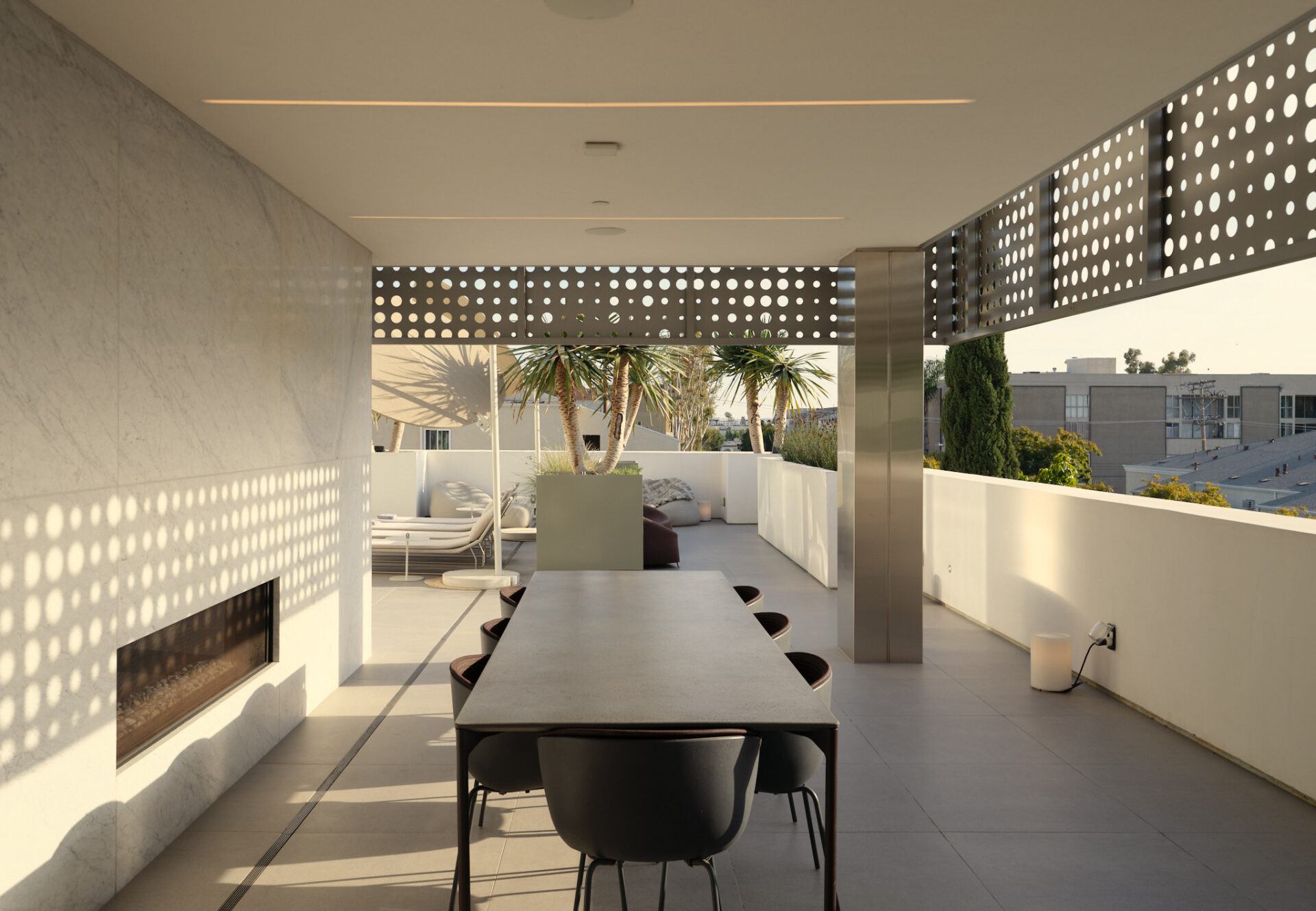 Rooftop sundeck dining area with modern table and chairs beneath perforated metal panels, lit by sunset glow.