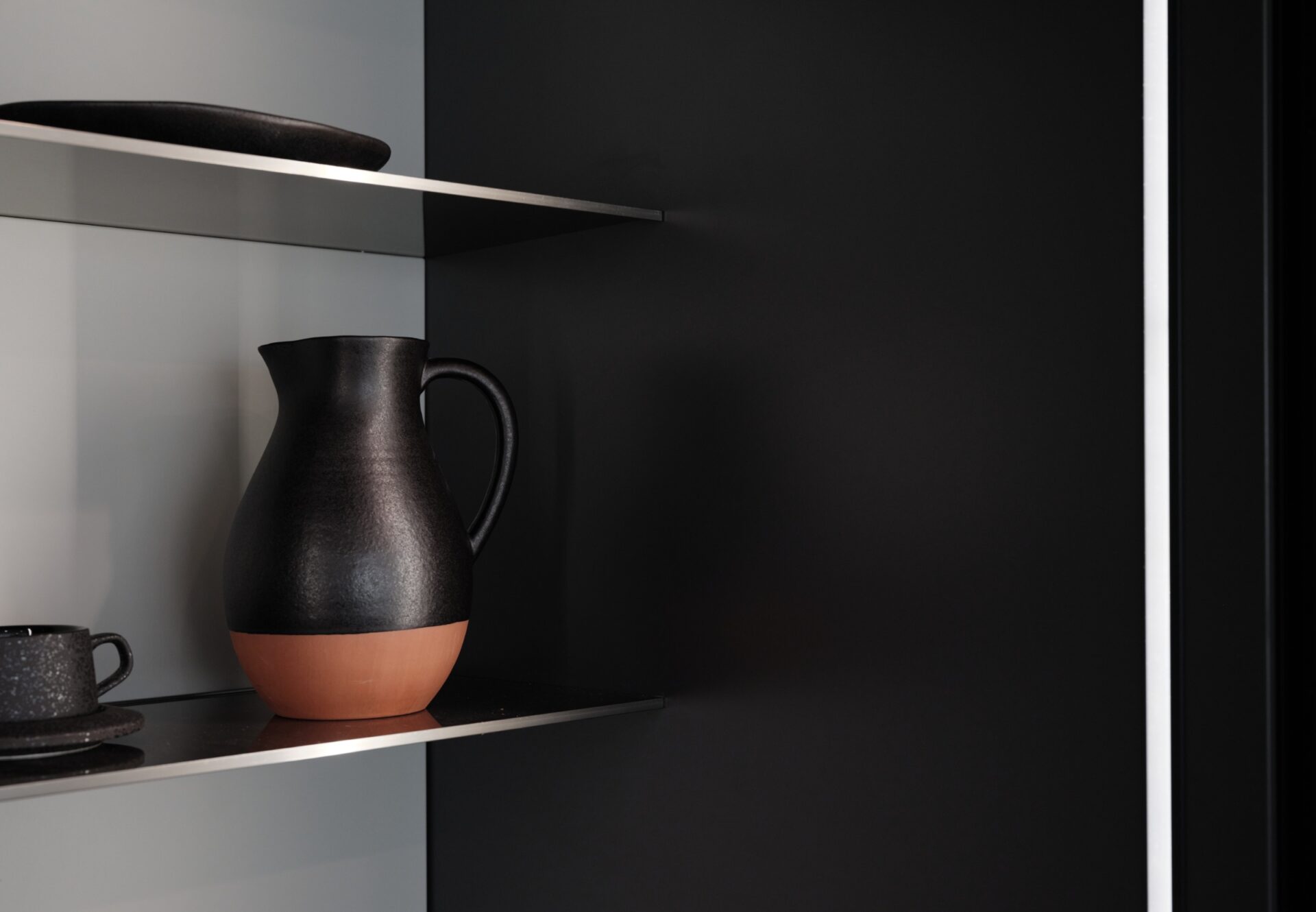Close-up of built-in kitchen shelf with matte black and terracotta pitcher, softly lit by recessed lighting against dark backdrop.