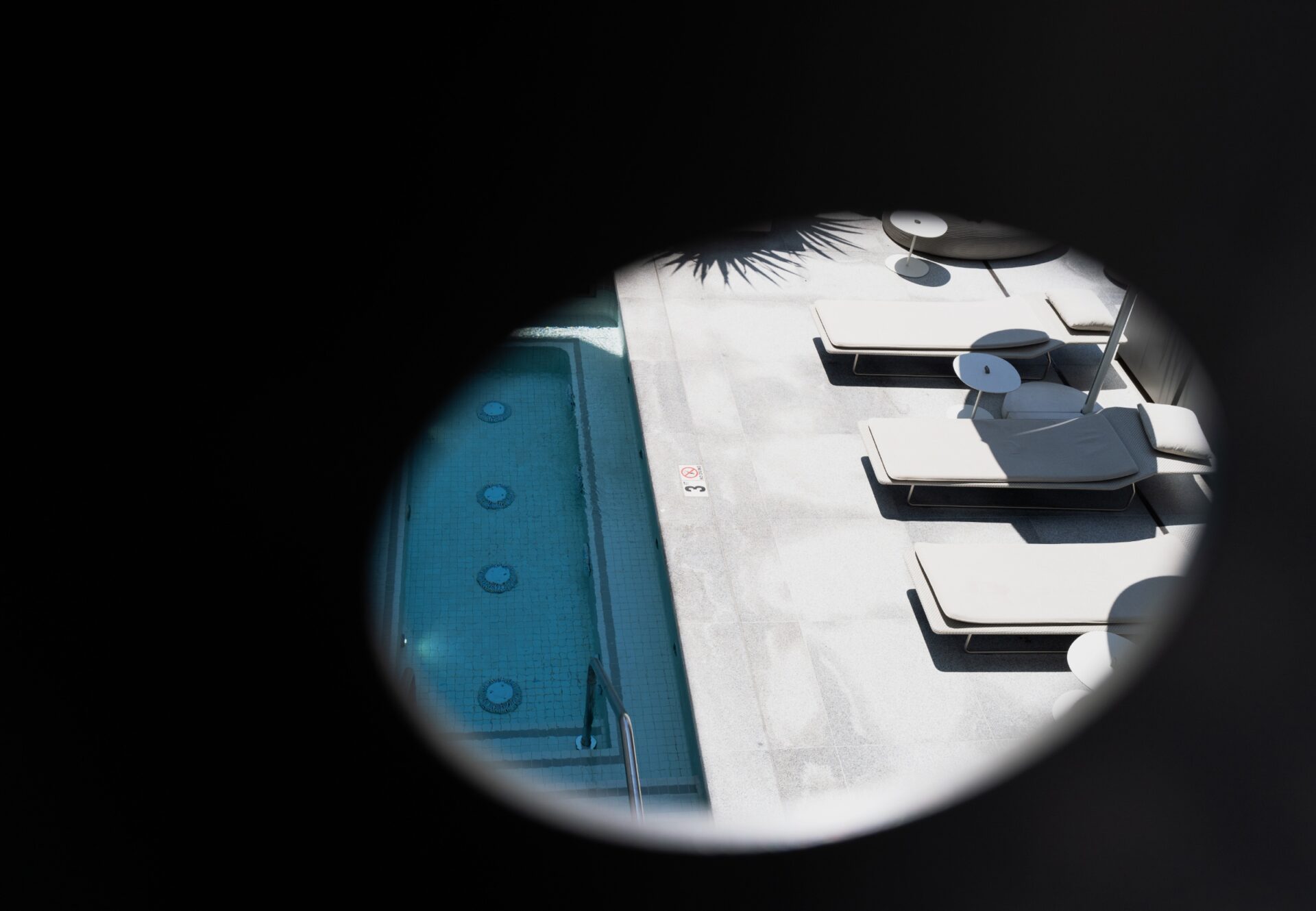 View of rooftop pool and loungers through a perforated steel screen, with a glimpse of the spa pool and sunlit sundeck below.