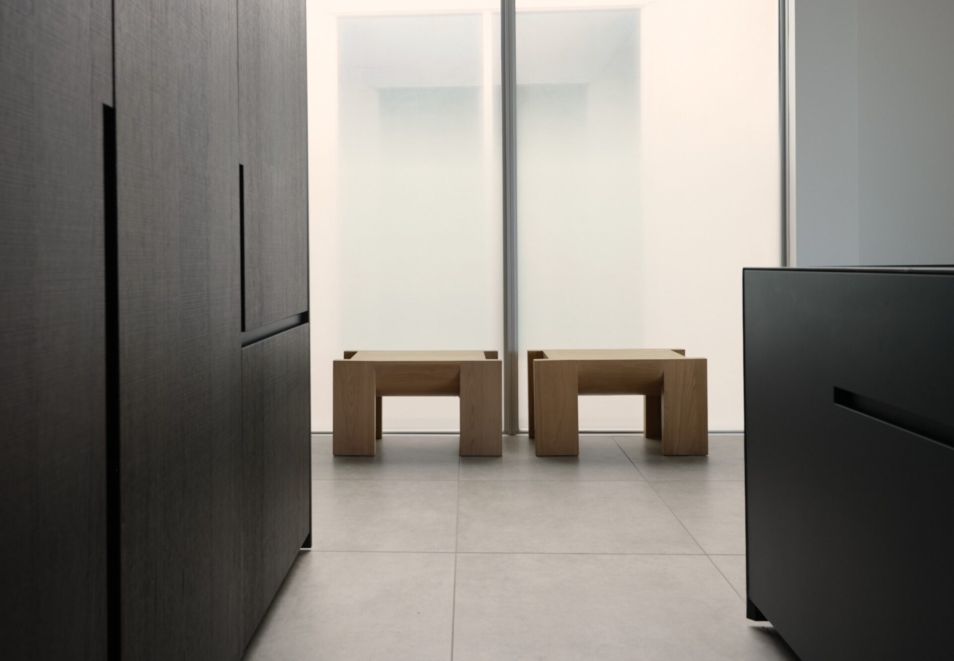 Minimalist kitchen with dark oak cabinetry, marble island, and sunlit atrium featuring frosted glass panels and wooden stools.
